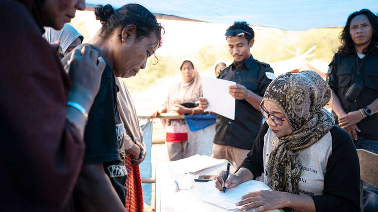 2019-nothilfe-tsunami-registrierung-verteilung-indonesien_C_acted.jpg Zwei Frauen stehen vor einem Tisch, dahinter sitzt eine weitere Frau mit einer Liste.