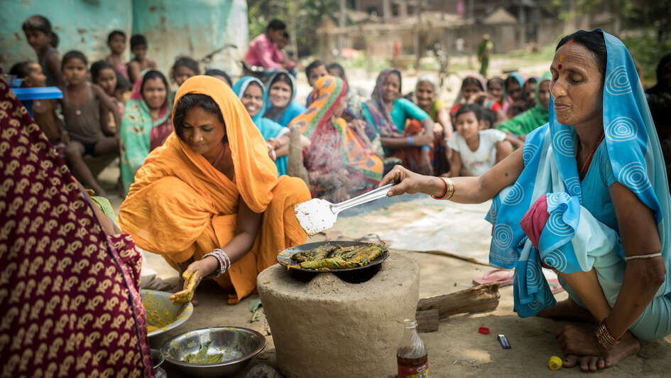 Zwei Frauen kochen, im Hintergrund schauen mehrere Frauen zu.