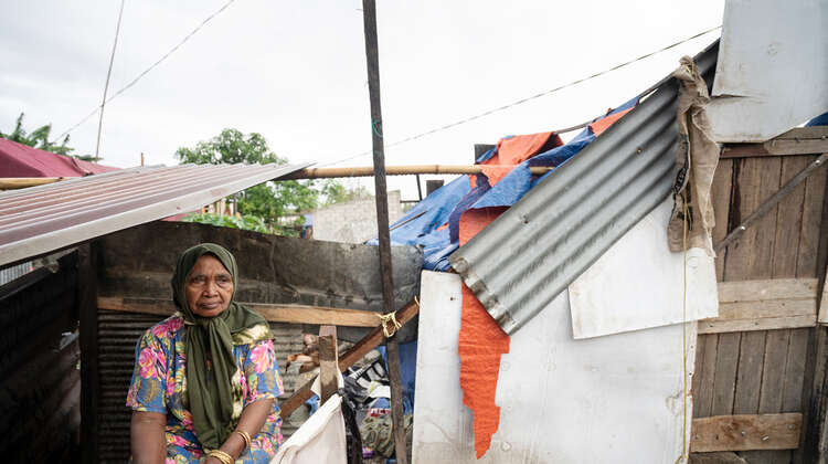 2019-nothilfe-tsunami-frau-notunterkunft-indonesien_C_acted.jpg Eine ältere Frau schaut traurig in die Ferne, hinter ihr sieht man ein provisorisches "Zelt" aus Planen und Wellblech.