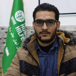 Portraitfoto von Welthungerhilfe-Mitarbeiter Adib Abokhors, der in Winterjacke vor einem Banner mit dem Logo der Welthungerhilfe sitzt