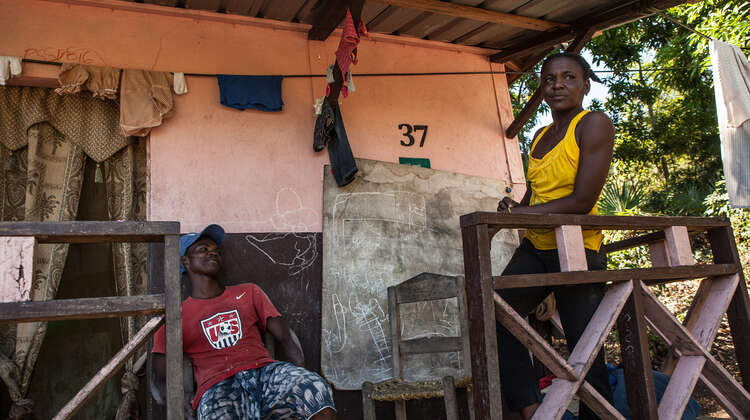 Eine Frau und ein Mann auf einer Veranda, die Frau steht am Geländer, der Mann sitzt an der Hauswand.