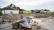 2019-nothilfe-tsunami-ueberlebende-abwasch-indonesien_C_acted.jpg Eine Frau sitht auf einem Stück Holz, vor ihr ein Wäschekübel. Um sie herum sieht man die Zerstörung, die der Tsunami angerichtet hat.
