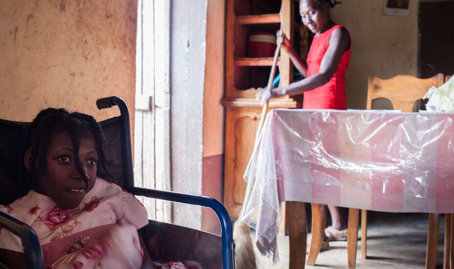 Im Hintergrund fegt Petit Lexi mit einem Besen. Im Vordergrund sitzt ihre Tochter Marie-Mikibelle in einem Rollstuhl.