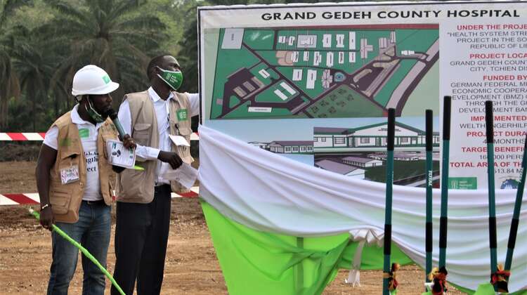Zwei Männer enthüllen eine Tafel mit dem Grundriss des geplanten Grand Gedeh County Hospital, Zwedru, Liberia, 2021 Zwei Männer enthüllen eine Tafel mit einem Bauplan.