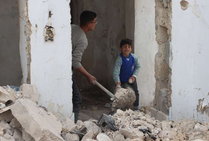 Ein älterer und ein jüngerer Junge räumen mit Schaufeln Schutt aus einem zerstörten Haus.