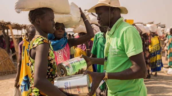Im Vordergrund: Eine Frau erhält von einem Mann in grünem T-shirt Nahrungsmittel. Im Hintergrund sieht man Menschen, die in einer Schlange anstehen.