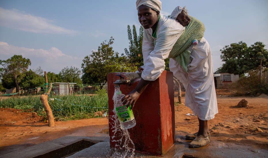 Eine Frau mit einem Baby auf dem Rücken holt Wasser an einem Brunnen.