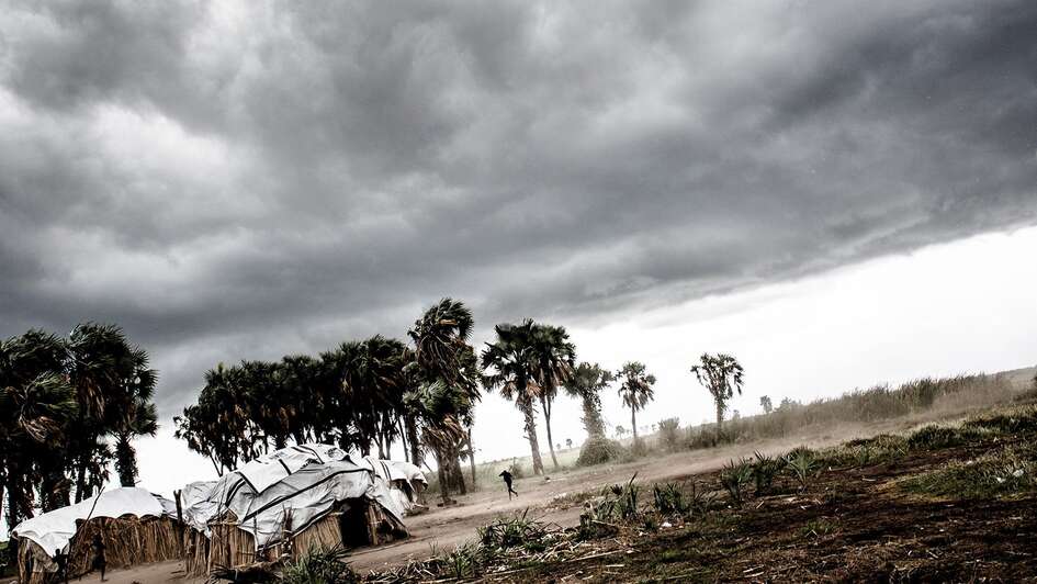 Dunkle Wolken am Himmel, neben aus Holz und Planen gebaute Häuser stehen Palmen, die sich im Wind beugen.
