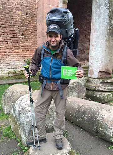 Ronny Lasner in Wanderkleidung und mit Rucksack hält das Logo der Welthungerhilfe in der Hand.