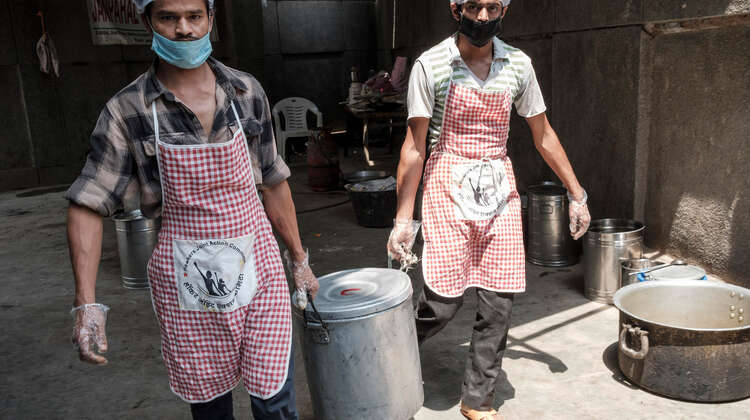 2020-coronavirus-slum-essen-kochen-ngo-mitarbeiter-delhi-indien-c-florian-lang.jpg Zwei Männer mit Schürze tragen einen großen Topf.
