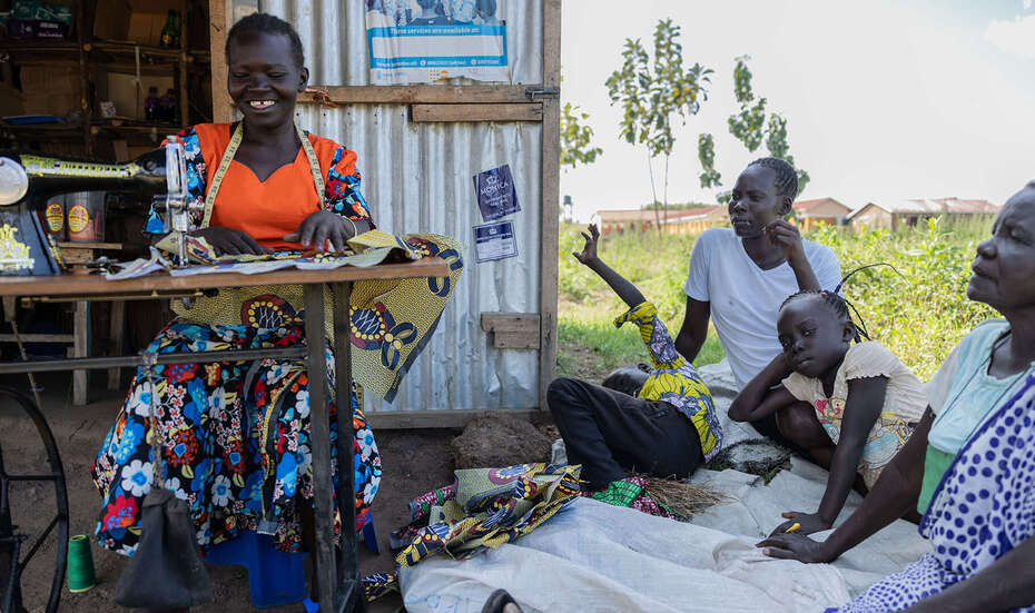 Frau arbeitet an Nähmaschine und lächelt, neben ihr sitzen mehrere Personen im Gespräch und schauen ihr zu