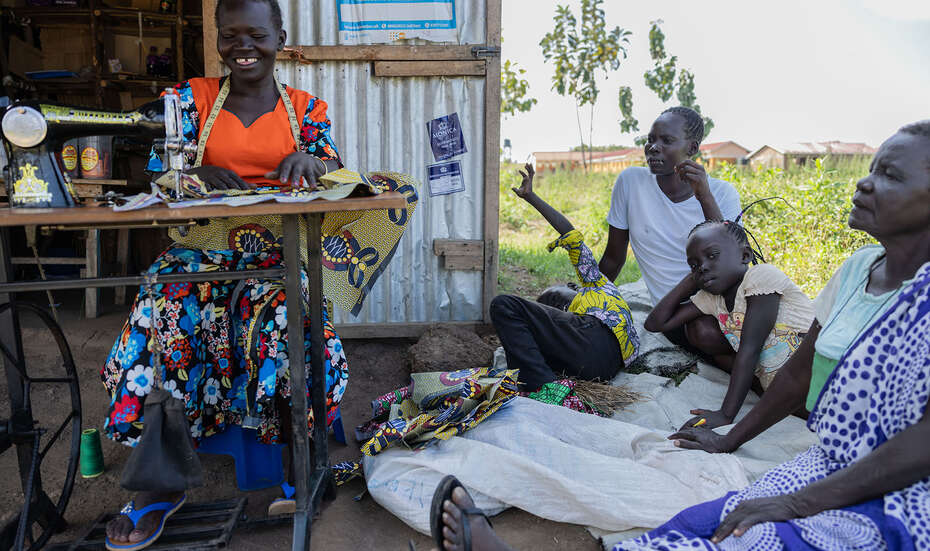 Frau arbeitet an Nähmaschine und lächelt, neben ihr sitzen mehrere Personen im Gespräch und schauen ihr zu