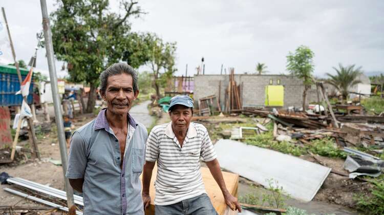 2019-nothilfe-tsunami-ueberlebende-zerstoerung_C_acted.jpg Zwei Männer schauen in die Kamera, hinter ihnen sieht man, was von den Häusern noch übrig geblieben ist.