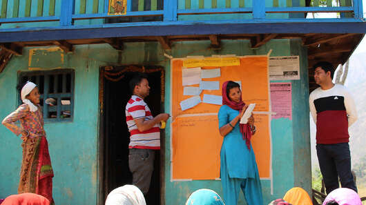 Mehrere Frauen sitzen auf dem Boden und schauen sich einen Vortrag an, weiter hinten stehen zwei Frauen und zwei Männer an einer Tafel.