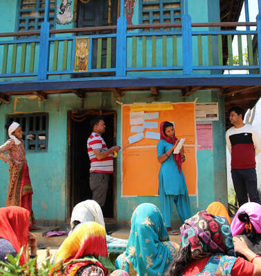 Mehrere Frauen sitzen auf dem Boden und schauen sich einen Vortrag an, weiter hinten stehen zwei Frauen und zwei Männer an einer Tafel.