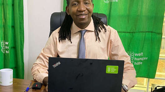 Ein Mann sitzt an einem Schreibtisch und lächelt in die Kamera.