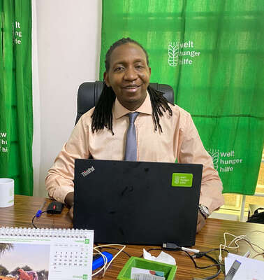 Ein Mann sitzt an einem Schreibtisch und lächelt in die Kamera.