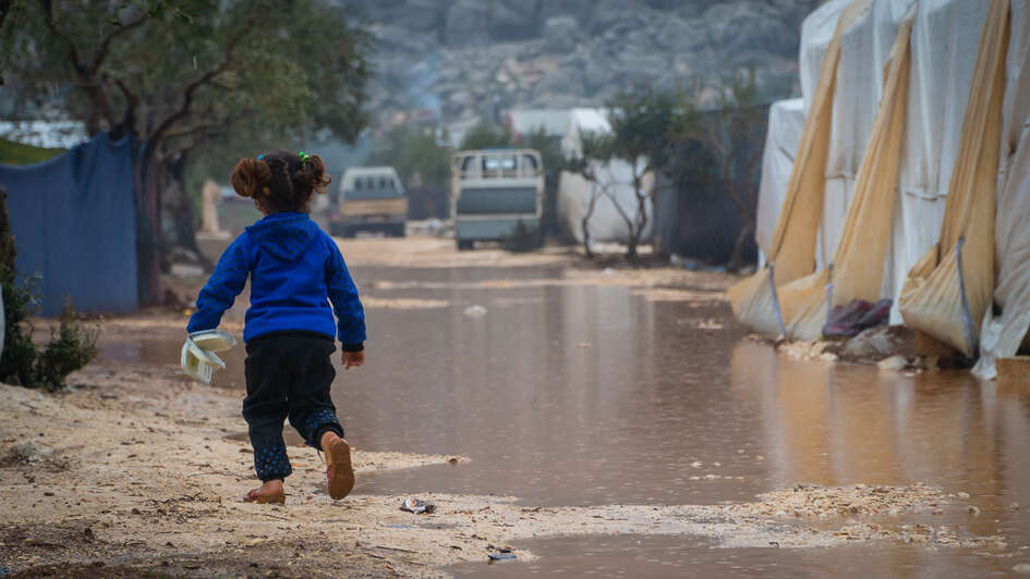 Ein kleines Mädchen von hinten, es läuft auf einem unbefestigten Weg, der überschwemmt ist. Im Hintergrund sieht man Zelte.