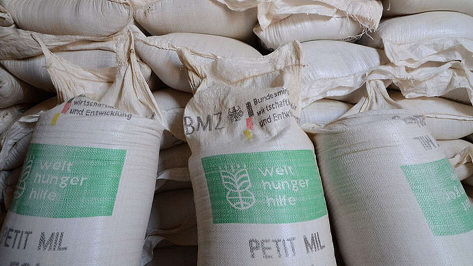 Drei vor Mehlsäcke der Welthungerhilfe stehen vor einem Sackstapel..