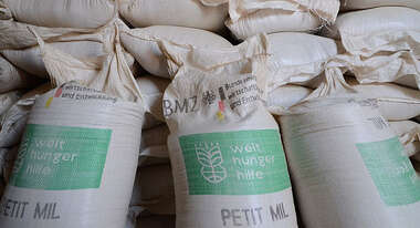 Drei vor Mehlsäcke der Welthungerhilfe stehen vor einem Sackstapel..