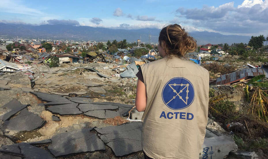 Eine Frau steht mit dem Rücken zur Kamera und schaut auf eine Reihe zerstörter Häuser.