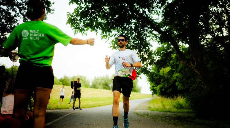 Eine Helferin in einem Welthungerhilfe-T-Shirt reicht einem Läufer einen Becher mit Wasser