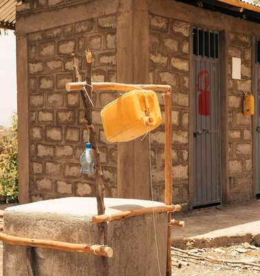 Im Vordergrund sieht man einen Brunnen, im Hintergrund ein Gebäude mit mehreren Türen.