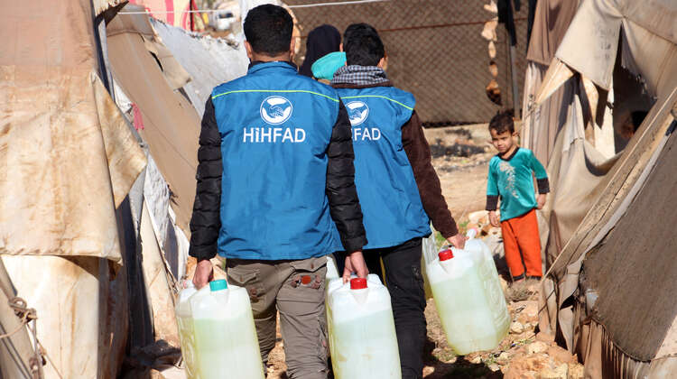 2020-winterhilfe-syrien-diesel-verteilung-hihfad-kind-welthungerhilfe.JPG HIHFAD-Mitarbeiter in blauen Westen tragen Diesel in Kunststoffkanistern zu einem Zelt. Ein Kind schaut ihnen entgegen.