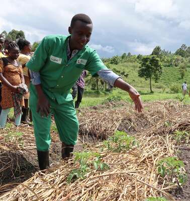 Ein Mann in grünem Overall zeigt jungen Menschen etwas auf einem Gemüsebeet.