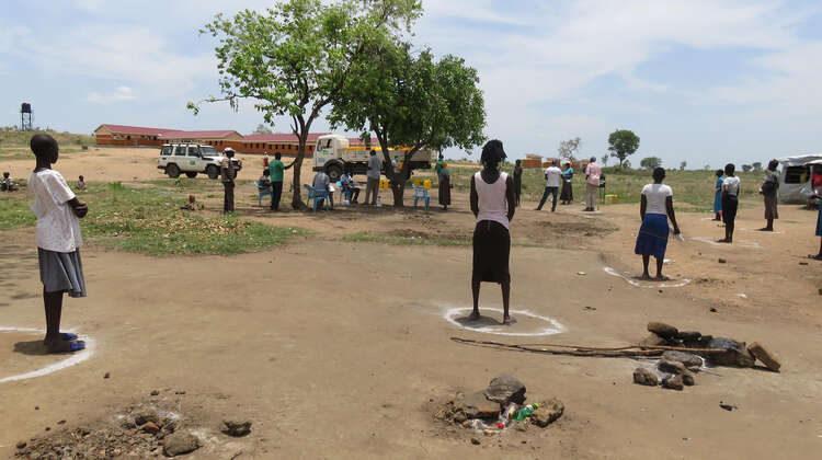 2020-corona-social-distancing-verteilung-fluechtlinge-uganda-c-welthungerhilfe.jpg Einige Menschen stehen mit dem Rücken zur Kamera in auf dem Boden aufgezeichnete Kreise.