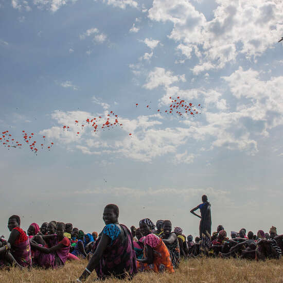 Auf dem Boden sitzen Menschen auf einem Feld, in der Ferne sieht man ein Flugzeug, das Nahrungsmittel aus der Luft ablädt.