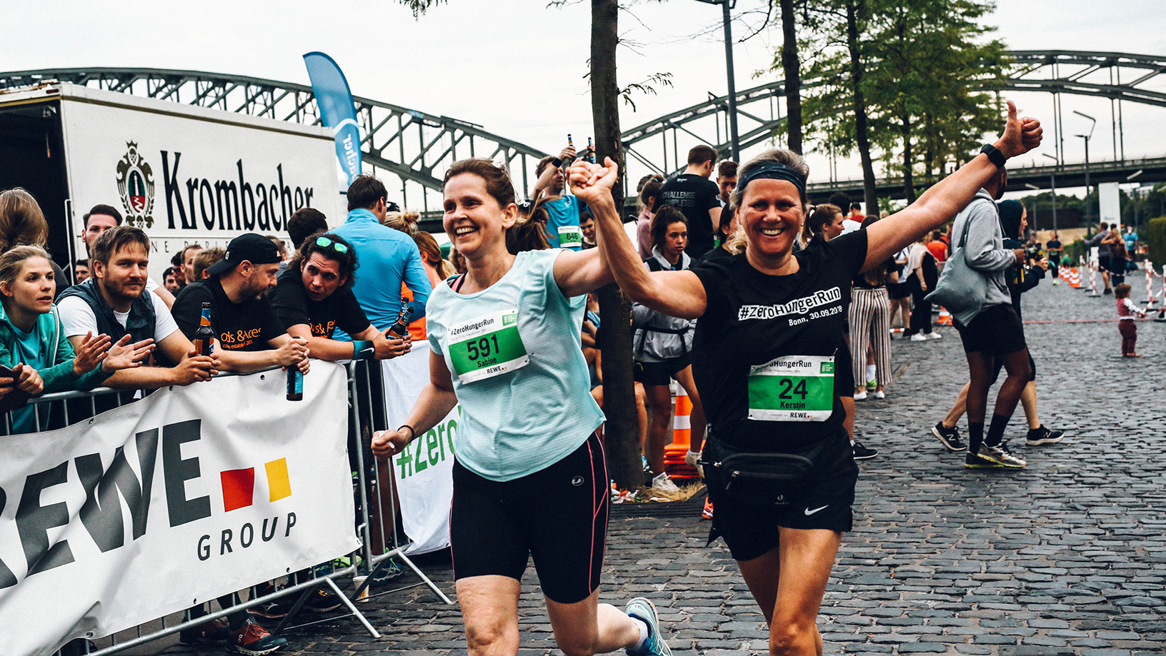 Zwei laufende Frauen in Sportkleidung heben jubelnd ihre Arme in die Höhe. Im Hintergrund sind Zuschauer und der Kölner Rheinauhafen zu sehen. 
