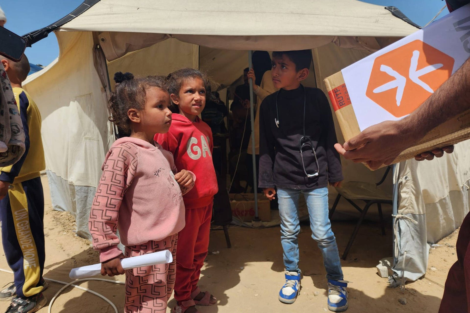 Kinder vor einem Zelt warten auf die Verteilung von Hilfsgütern, von rechts aus dem Bild kommt eine Person mit einer Kiste