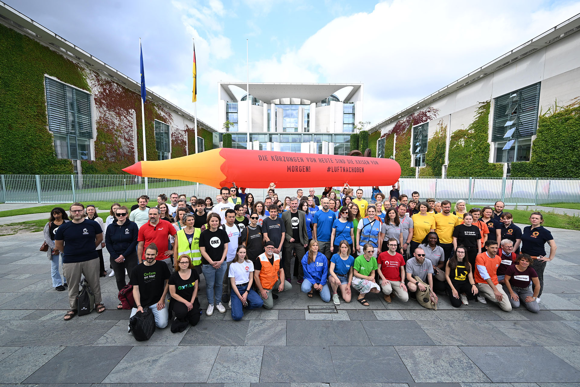 Gruppenfoto von demonstrierenden Personen von verschiedenen Hilfsorganisationen. Im Hintergrund das Kanzleramt und ein großer aufblasbarer Rotstift.