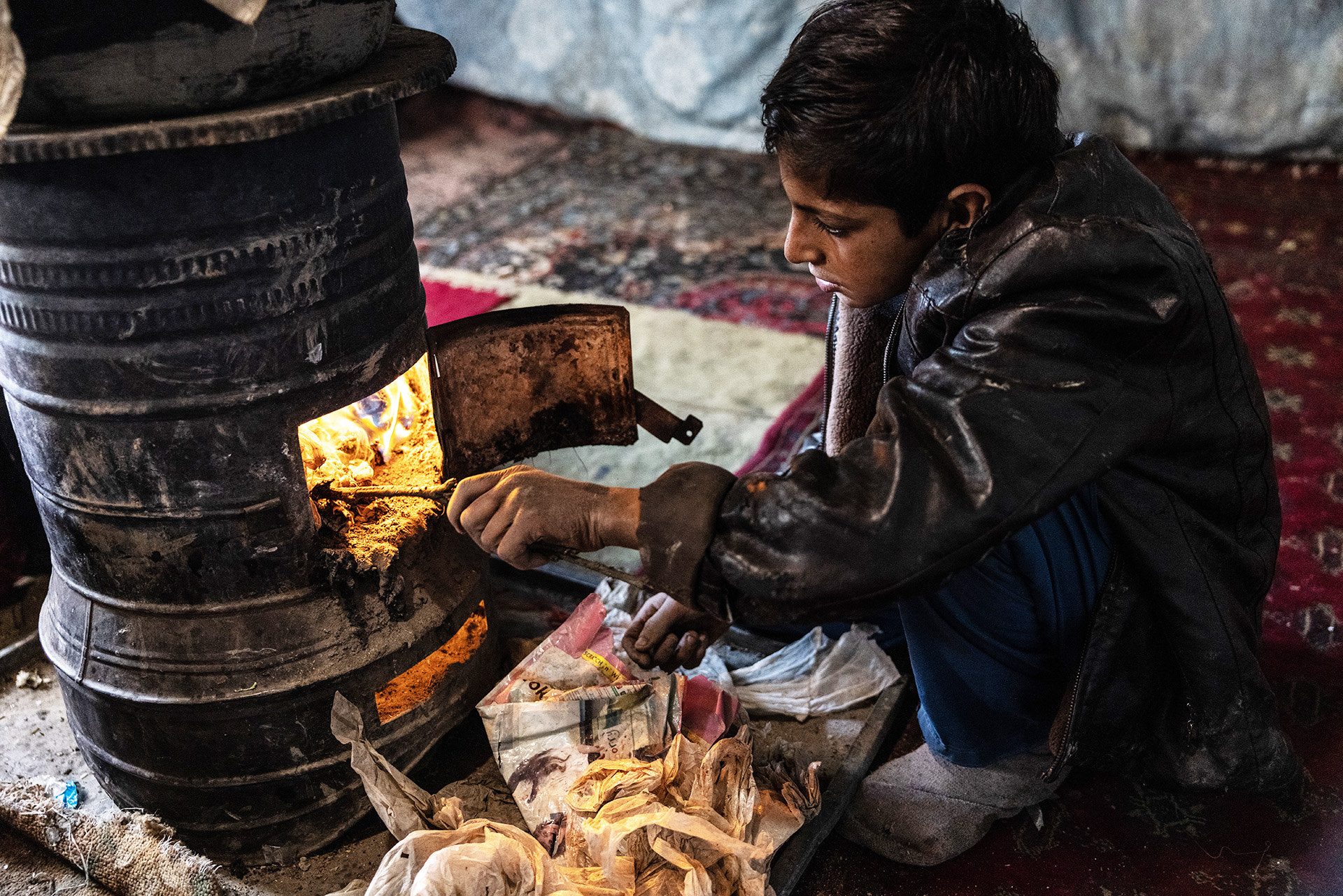 Ein Junge sitzt an einem Ofen und stochert mit einem Ast im Feuer.