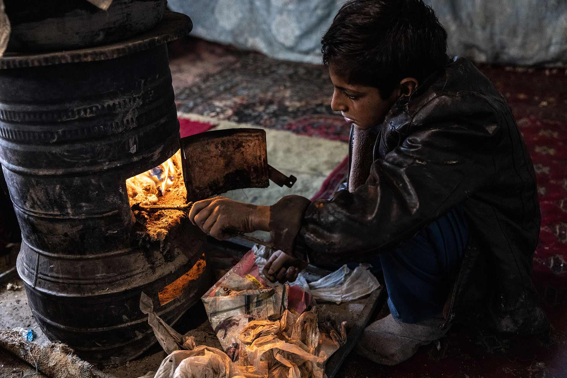 Ein Junge sitzt an einem Ofen und stochert mit einem Ast im Feuer.