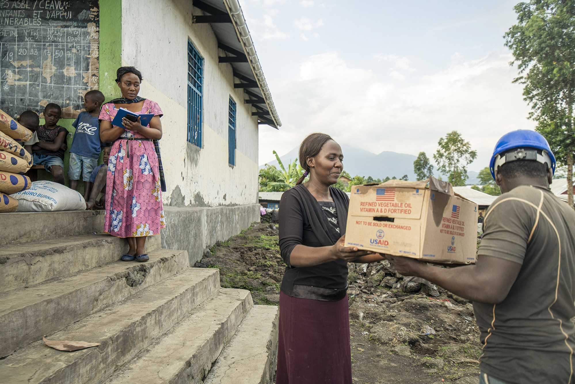 Ein Mann mit blauem Schutzhelm übergibt einer Frau mit schwarzem Shirt und weinrotem Rock vor einem Haus einen Pappkarton, in dem sich - so ist aufgedruckt - Hilfsgüter befinden. Eine Frau in rotem Kleid und mehrere Kinder stehen in einem Hauseingang im Hintergrund und schauen zu.