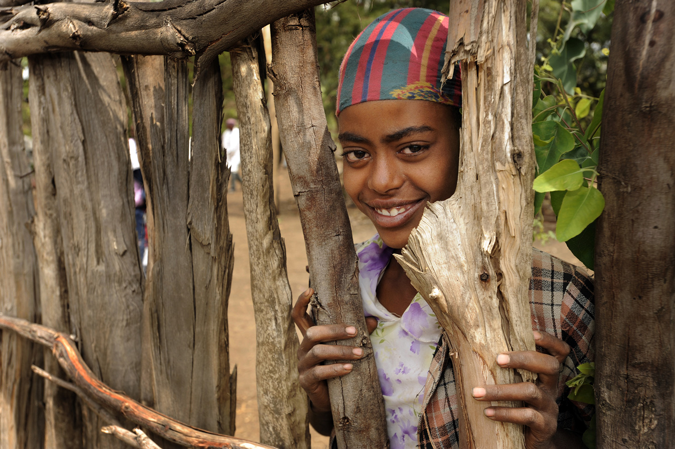 Kind schaut zwischen Baumstämmen hindurch und lacht in die Kamera.
