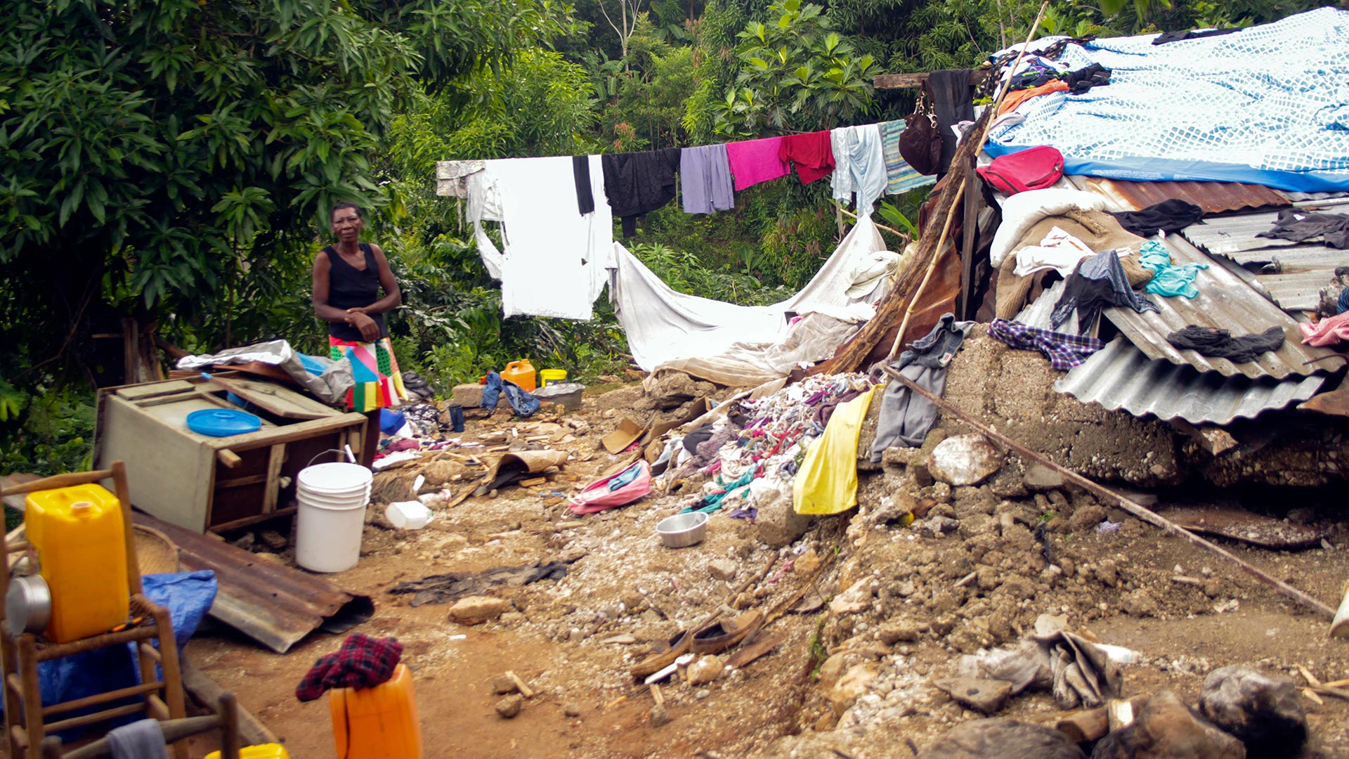 Eine Frau steht inmitten von Schutt und Teilen eines zerstörten Hauses. Im Hintergrund ist Wäsche aufgehängt. 