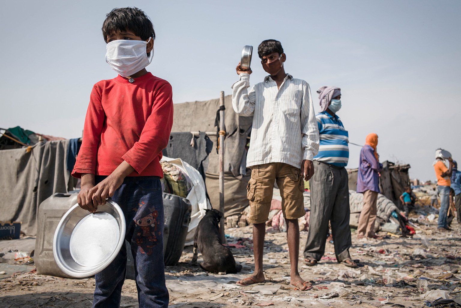 Mehrere Menschen, darunter auch Kinder, mit Mundschutz stehen in einer Schlange. Sie alle haben eine Metall-Schüssel in der Hand.