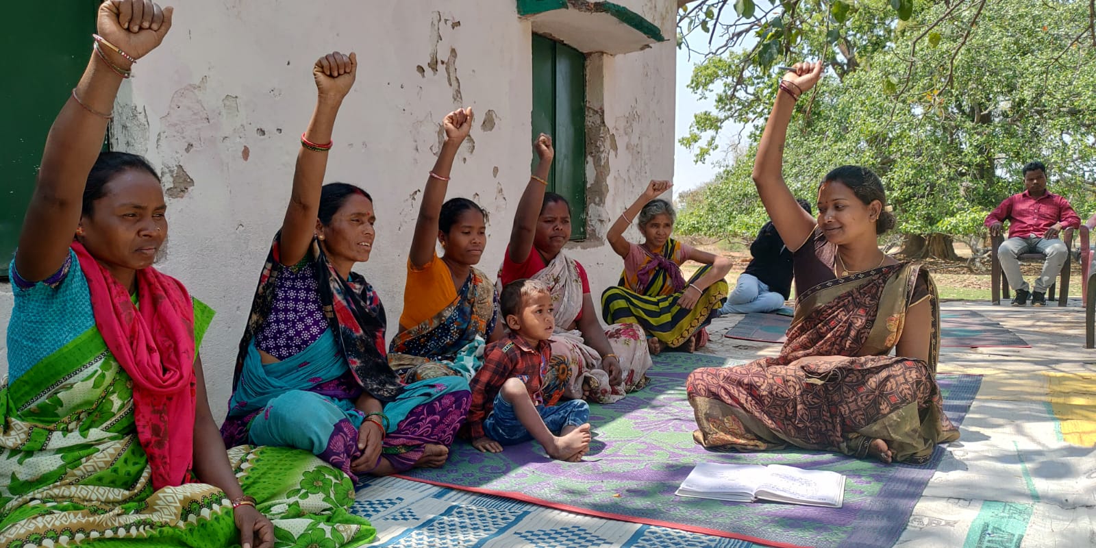 Frauen in Indien, draußen auf dem Boden sitzend bei einem Beratungsangebot. Sie strecken die Faust Richtung Himmel.