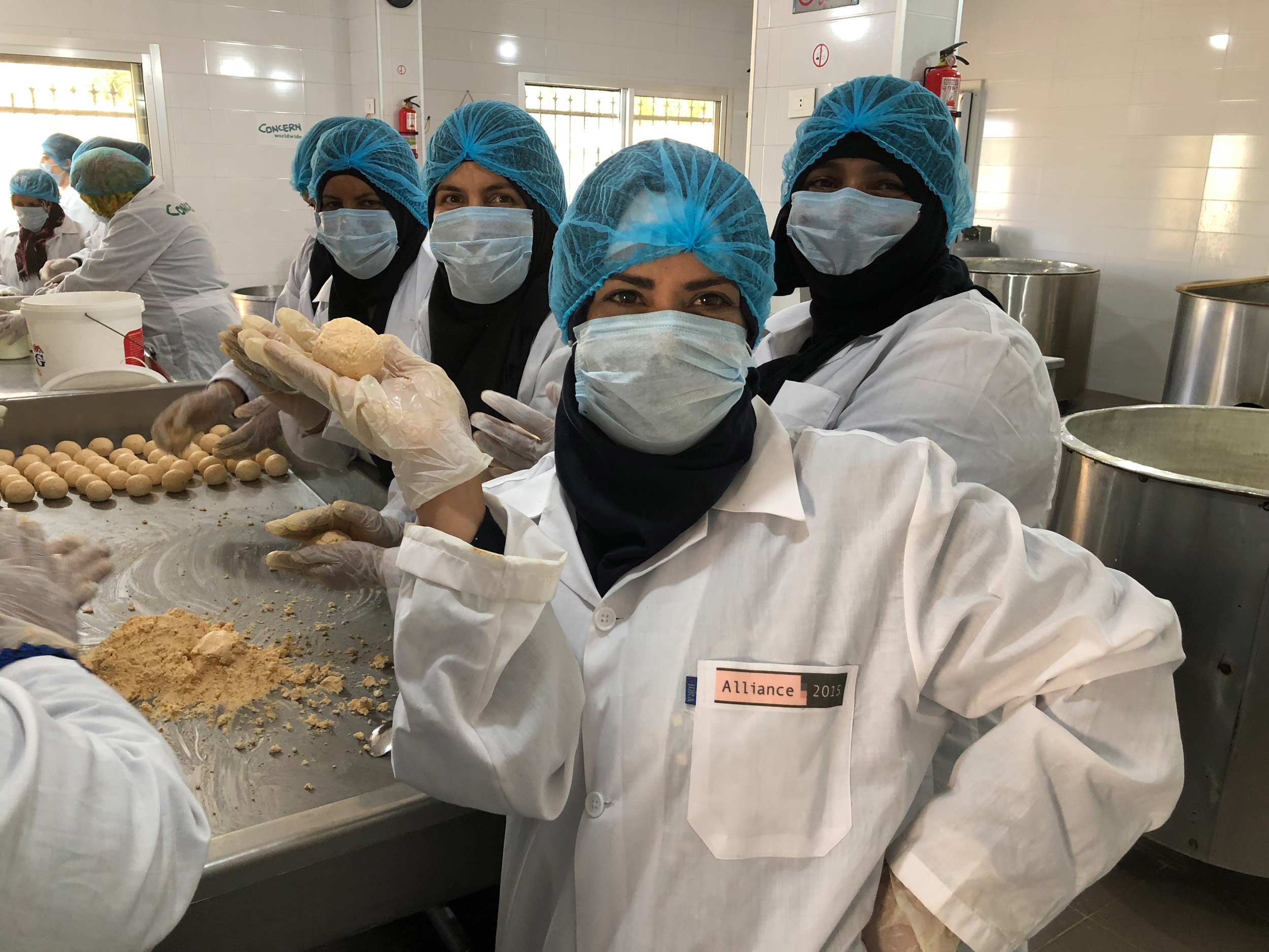 Eine Frau in weißem Kittel, blauer Haarhaube und Mundschutz hält ein Stück Käse hoch.