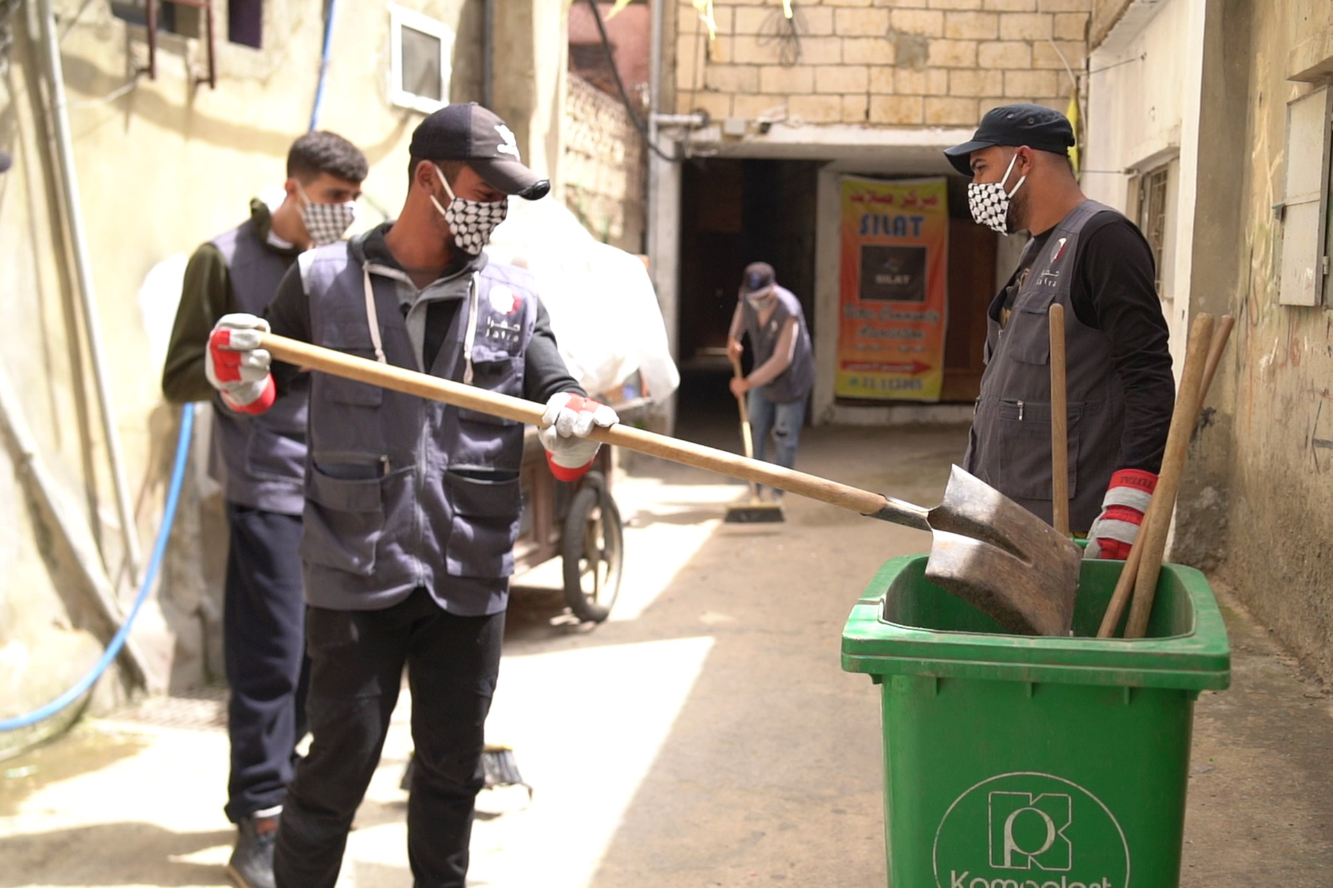 Drei Männer bei der Müllbeseitigung: Ein Mann wirft mit einer großen Schaufel etwas in eine grüne Tonne.