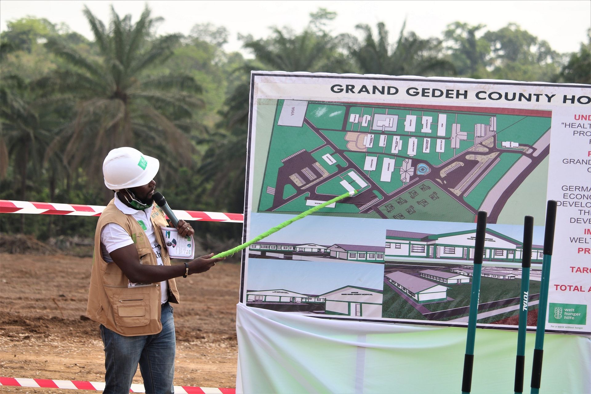 Ein Mann weist mit einem Zeigestock auf eine Tafel mit einem Bauplan.