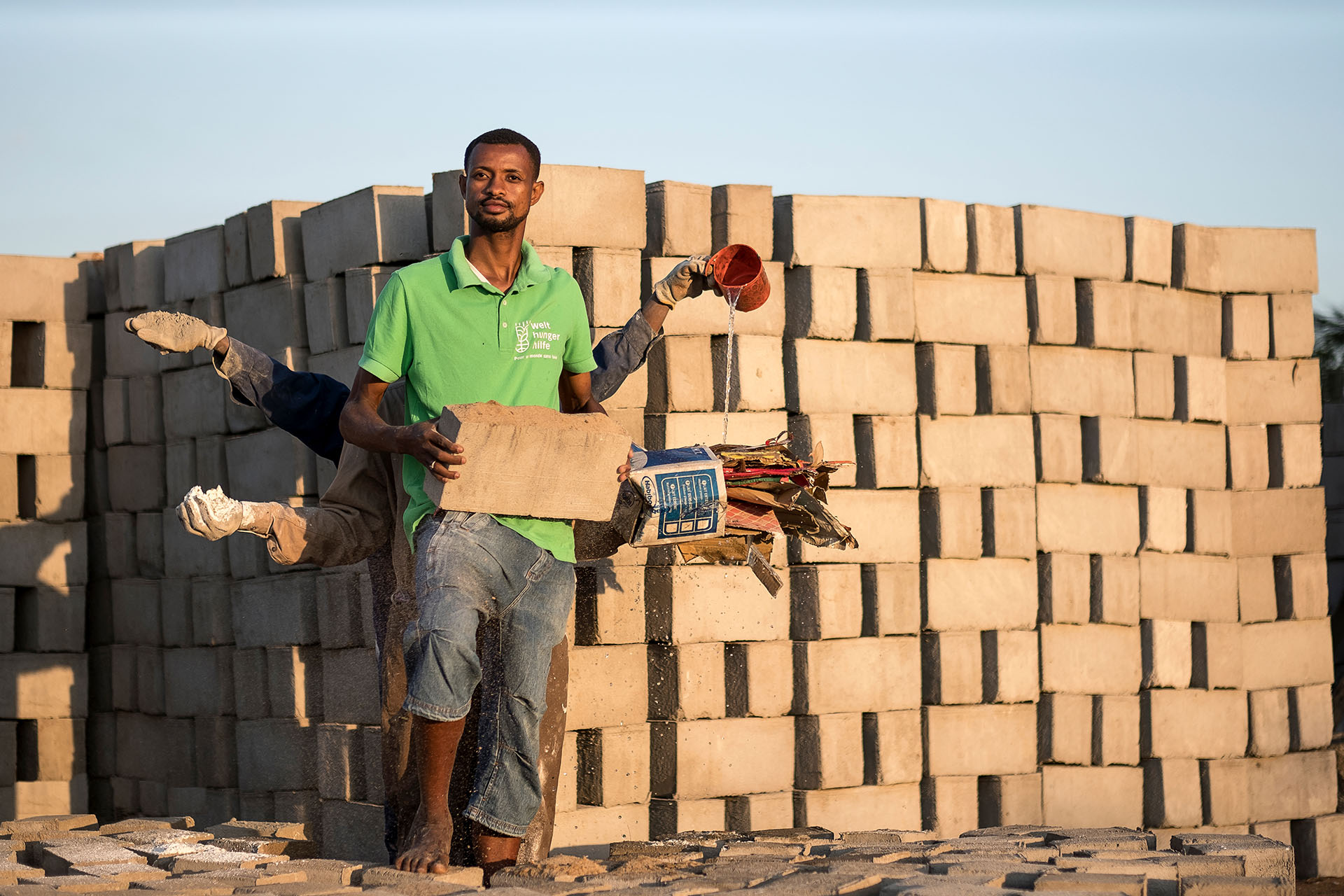 Drei Männer stehen hintereinander und strecken die Arme aus, in jeder Hand halten sie einen Rohstoff zur Herstellung von Ziegeln aus Abfallstoffen.
