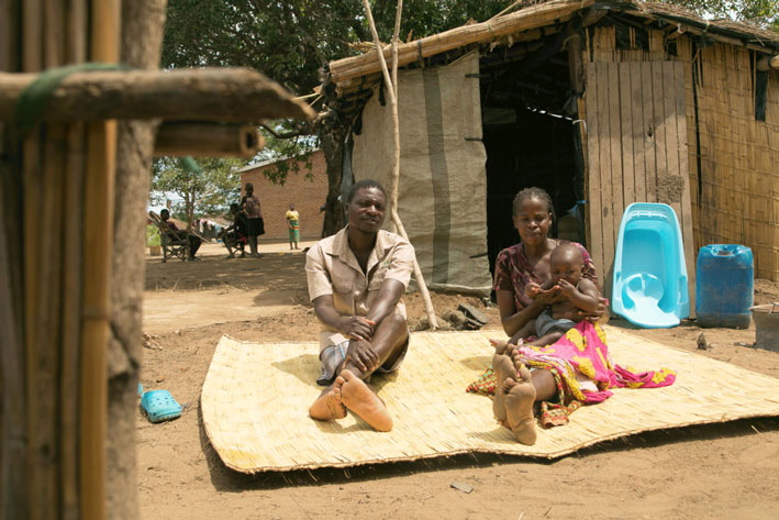 Auf einer Bastmatte sitzt ein Mann und eine Frau, die Frau hat ein Baby auf ihrem Schoß.