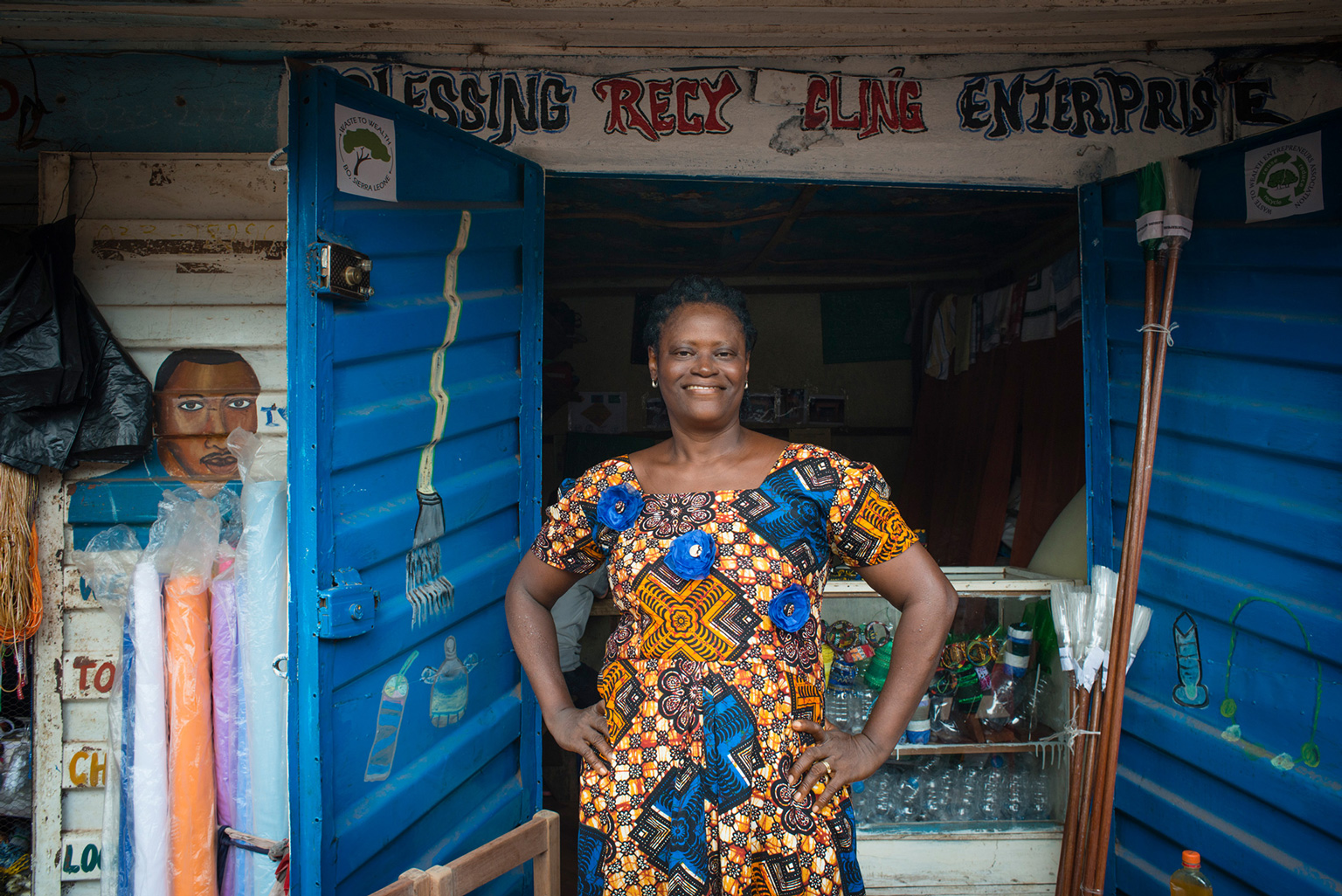 Eine Frau steht mit den Händen in den Hüften vor einem kleinen Laden, sie schaut lächelnd in die Kamera.