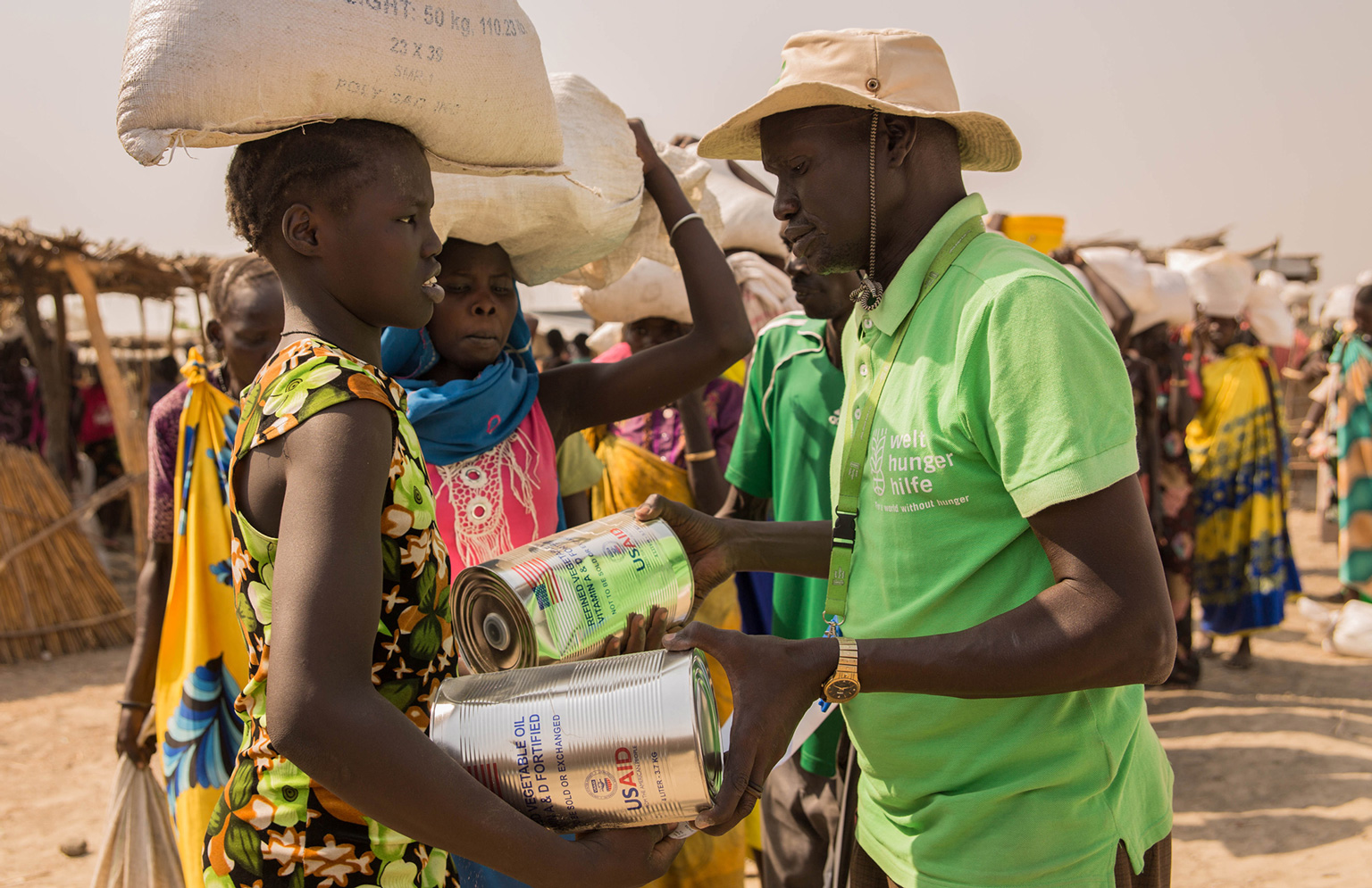 Im Vordergrund: Eine Frau erhält von einem Mann in grünem T-shirt Nahrungsmittel. Im Hintergrund sieht man Menschen, die in einer Schlange anstehen.
