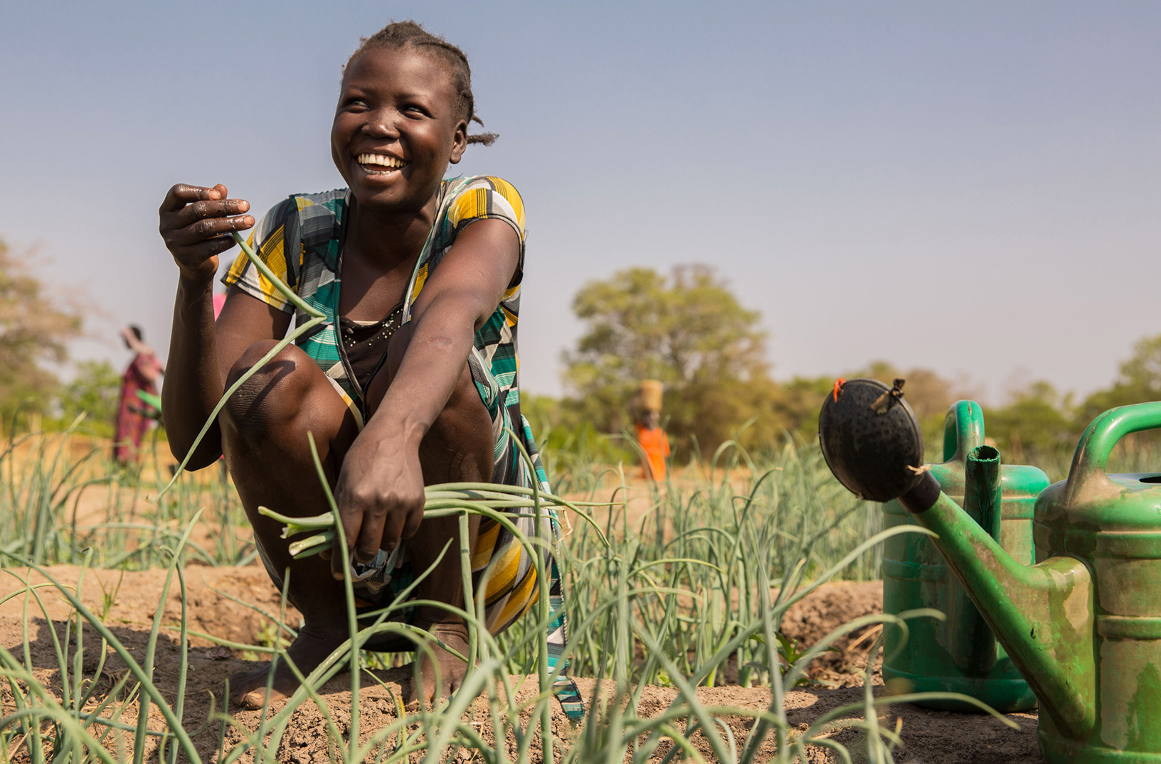 Eine Frau kniet lachend auf einem Gemüsebeet und hält eine Pflanze in der Hand.