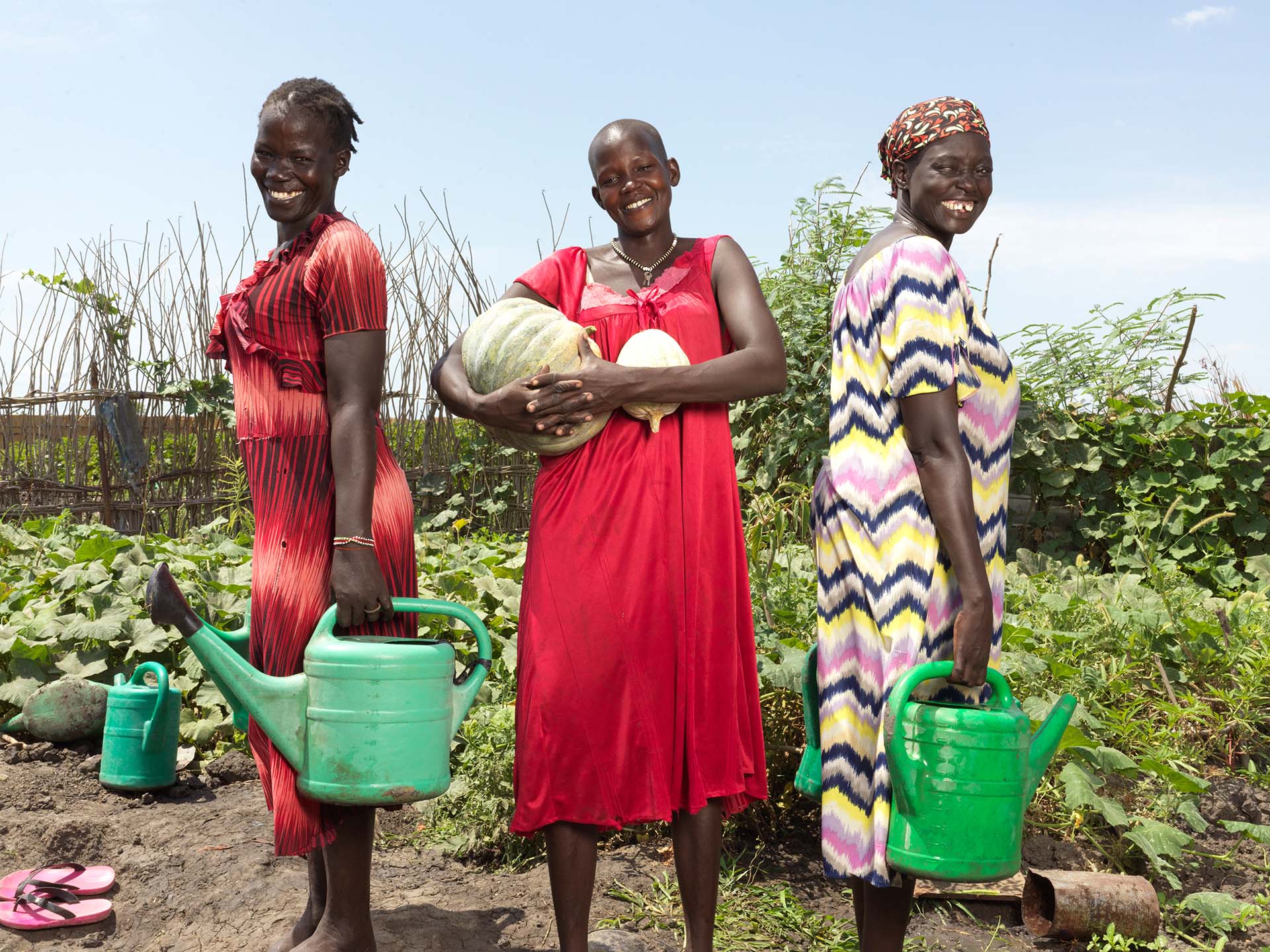 Drei Frauen in einem landwirtschaftlichen Garten halten Gießkannen und lächeln in die Kamera.